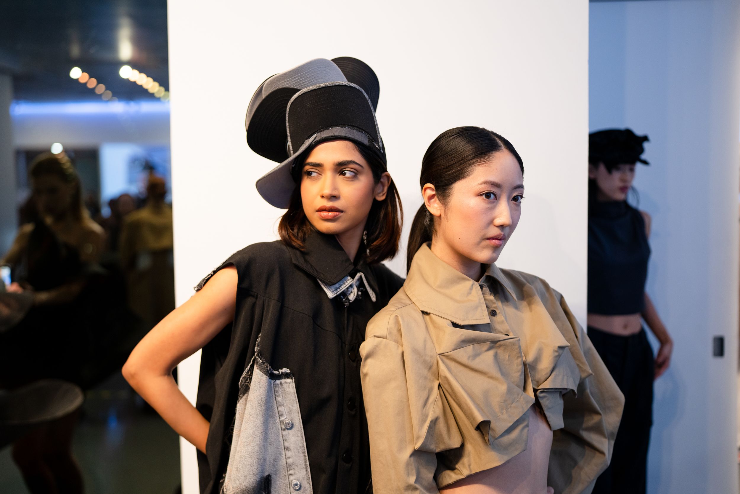 Two models pose backstage at a fashion show wearing avant-garde designs including a black hat and beige ruffled jacket.
