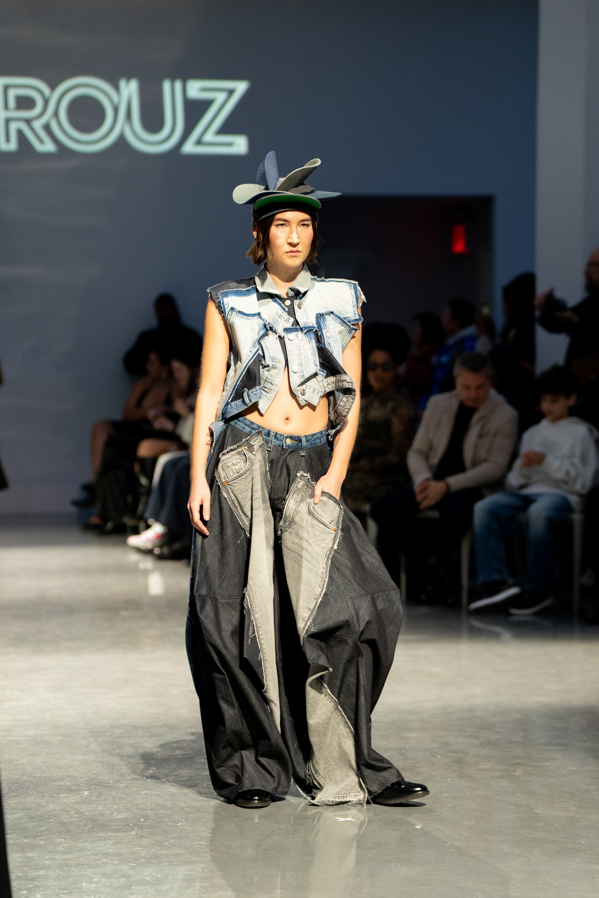 Model walks runway in oversized denim patchwork outfit with wide-leg pants and avant-garde hat at ROUZ fashion show.
