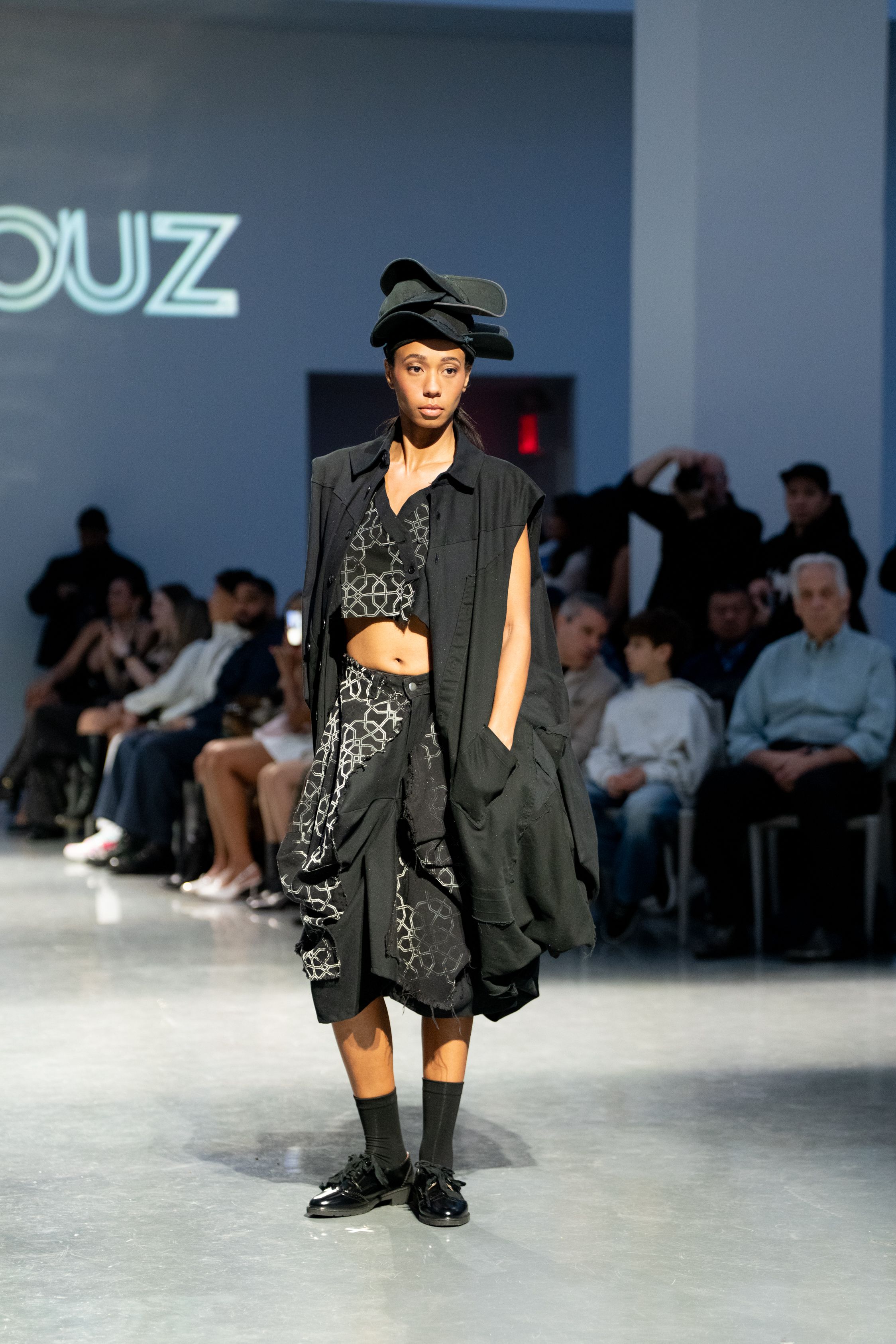 Model walks runway in dark avant-garde ensemble with layered skirt, vest, and black hat at fashion show.