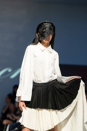 Model walks runway in white button-up shirt and layered black and white pleated skirt wearing decorative black mask.