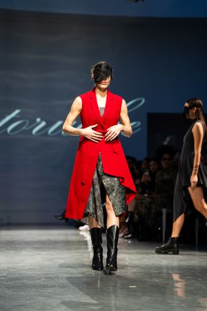 Model walks runway wearing bold red sleeveless longline vest over floral skirt with black knee-high boots and mask.