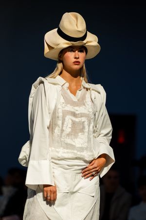 Female model on runway wearing white outfit with blazer, sheer top, skirt, and cream wide-brim hat with black band.