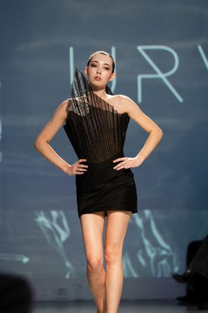Model walks runway in black one-shoulder mini dress with structured pleated bodice at fashion show.