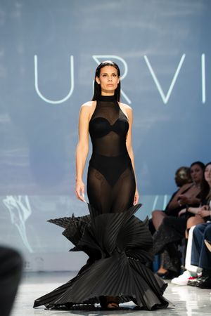 Model walks runway in sheer black halter gown with dramatic pleated skirt at URVI fashion show.