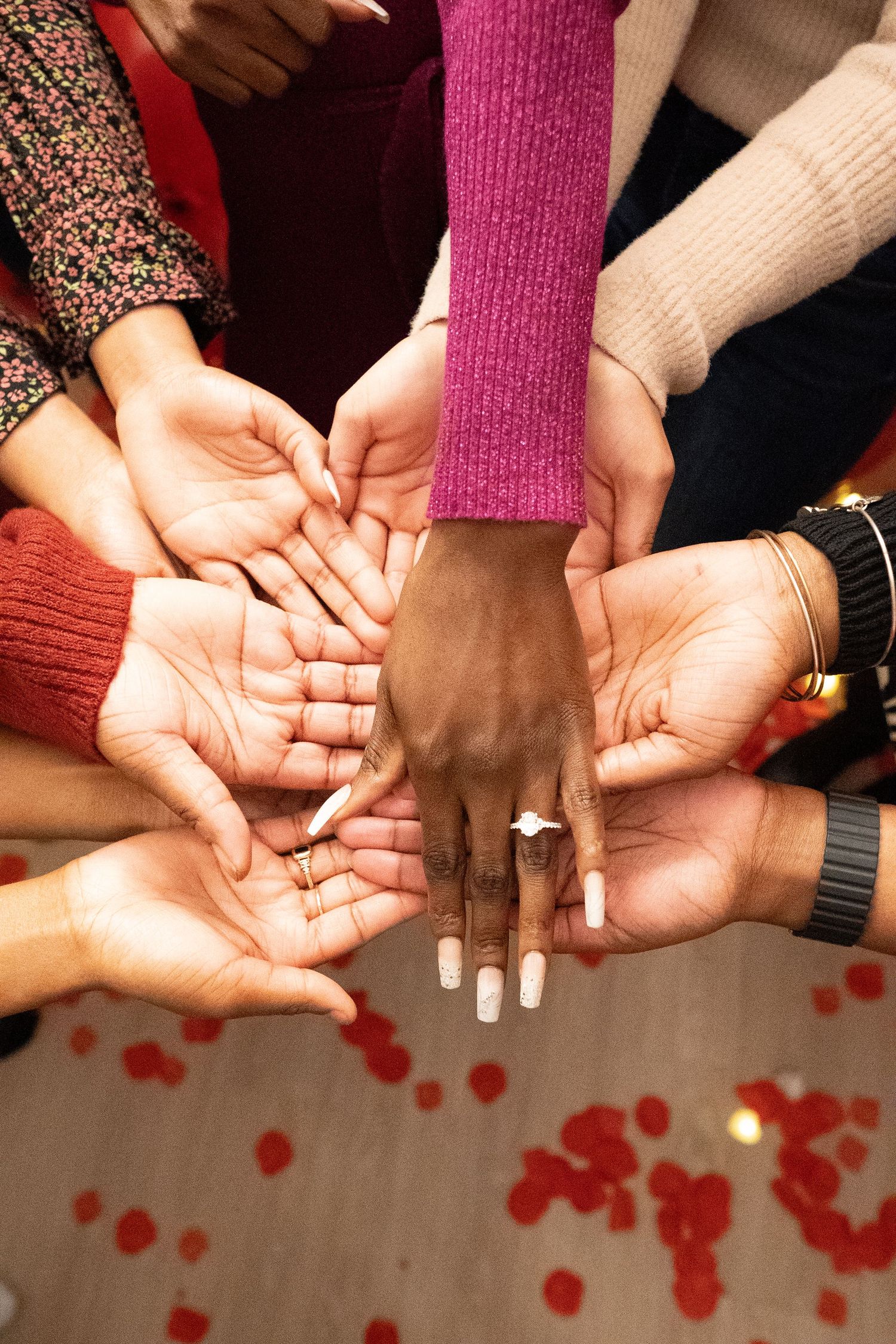 Multiple hands join together in a circle formation with red rose petals scattered below on a light surface.