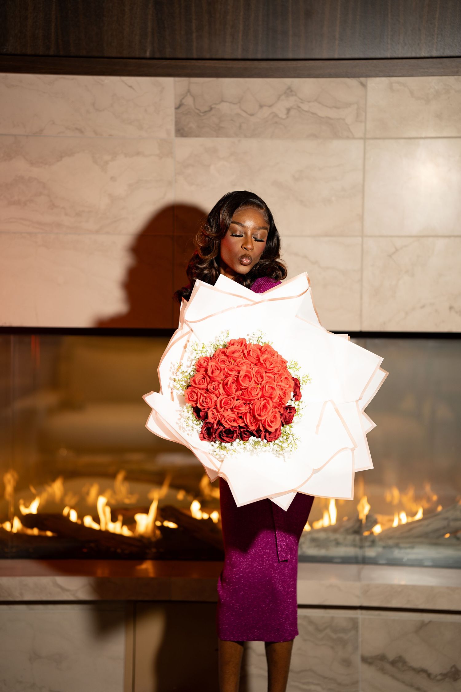 A person in a purple dress holds a large bouquet of red roses in front of a fireplace display.