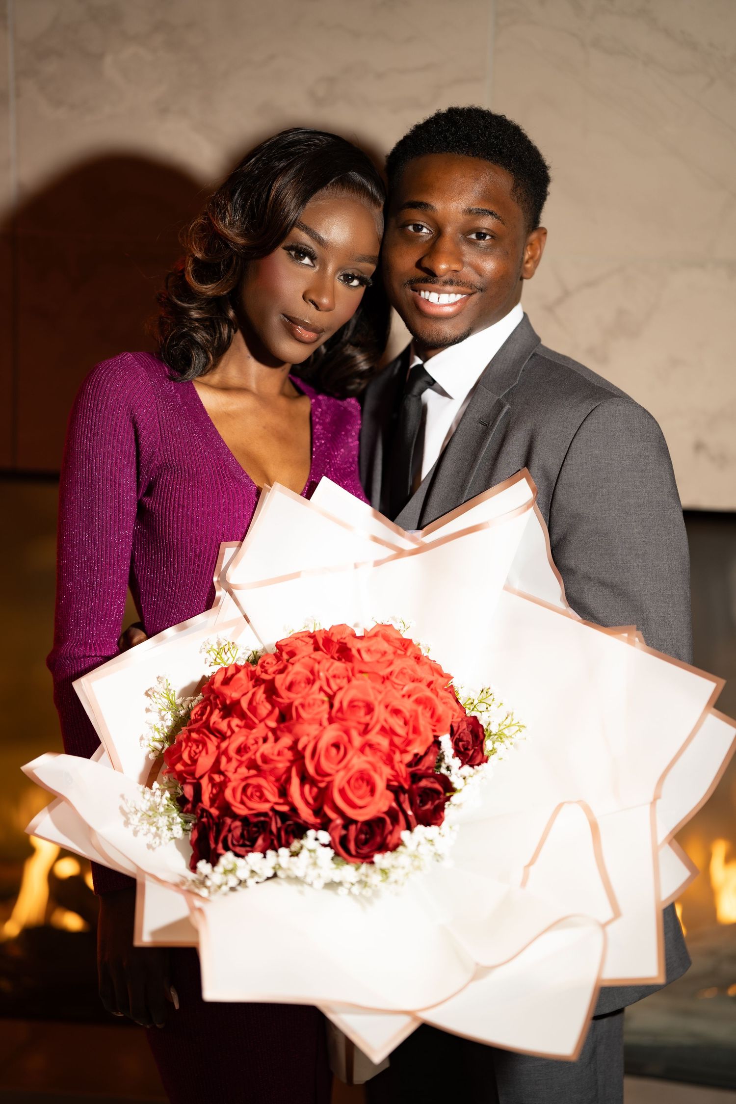 A couple poses with a beautiful bouquet of red roses wrapped in white paper at an elegant evening event.