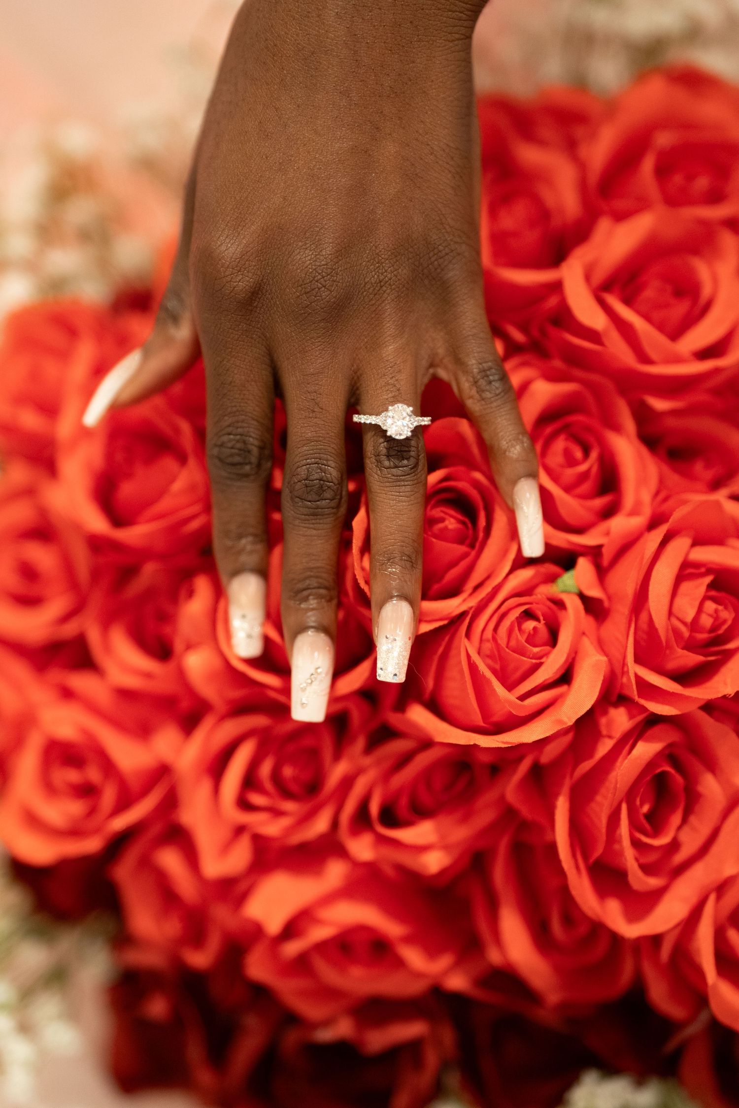 A hand wearing a diamond engagement ring rests delicately on a bed of vibrant coral roses.