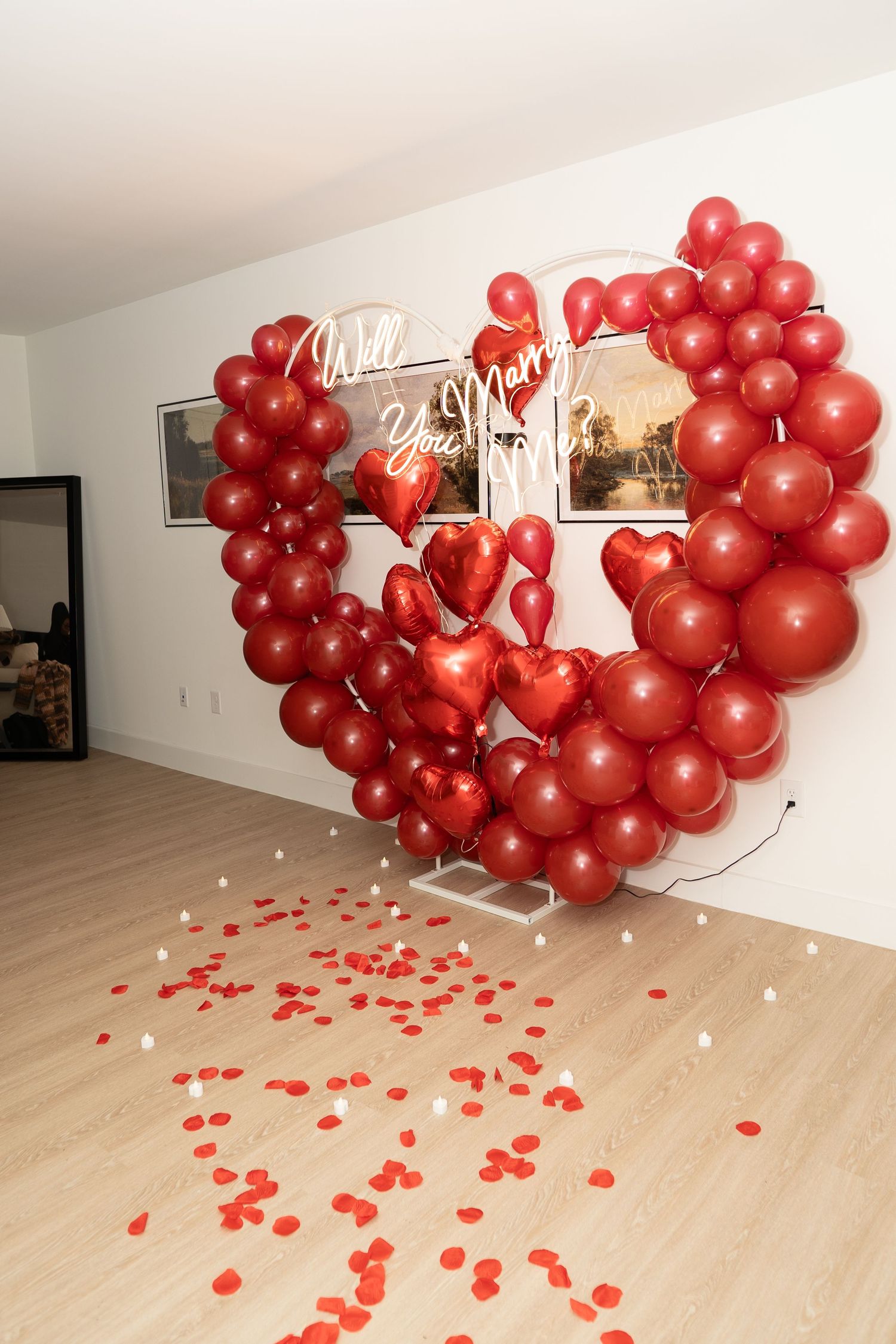 A romantic display of red balloons arranged in a heart shape with scattered rose petals on a wooden floor.