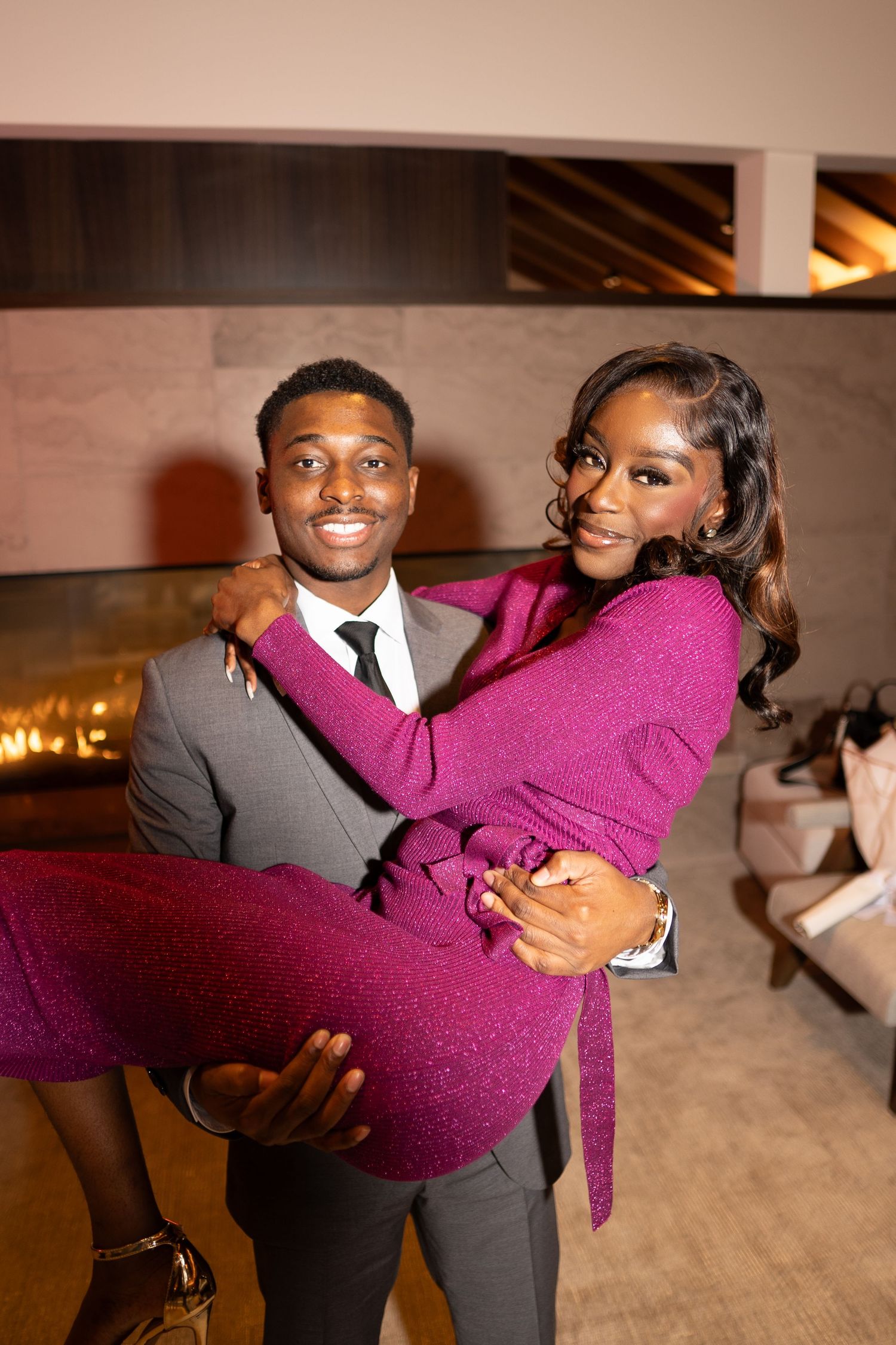 A couple in formal attire share a joyful moment as he carries her in a burgundy dress at an indoor venue.