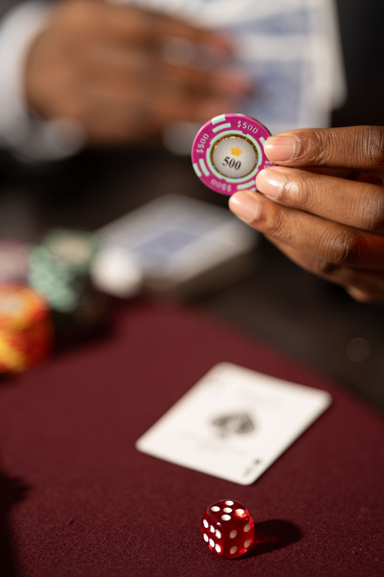 Hand holding a pink casino chip over a red gaming table with dice and playing cards visible in the background.