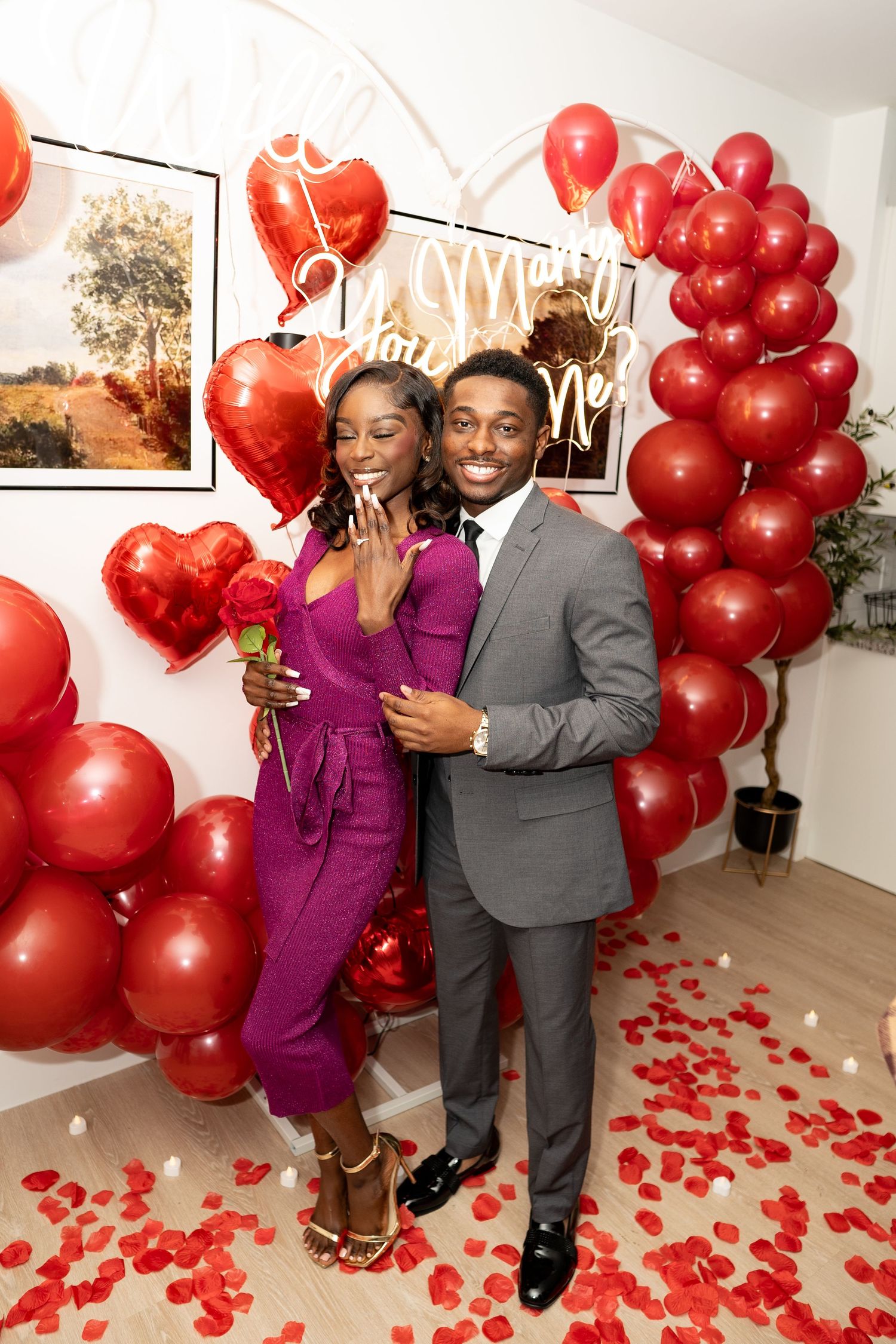 A couple celebrates in a room decorated with red balloons and rose petals for a romantic proposal or special occasion.