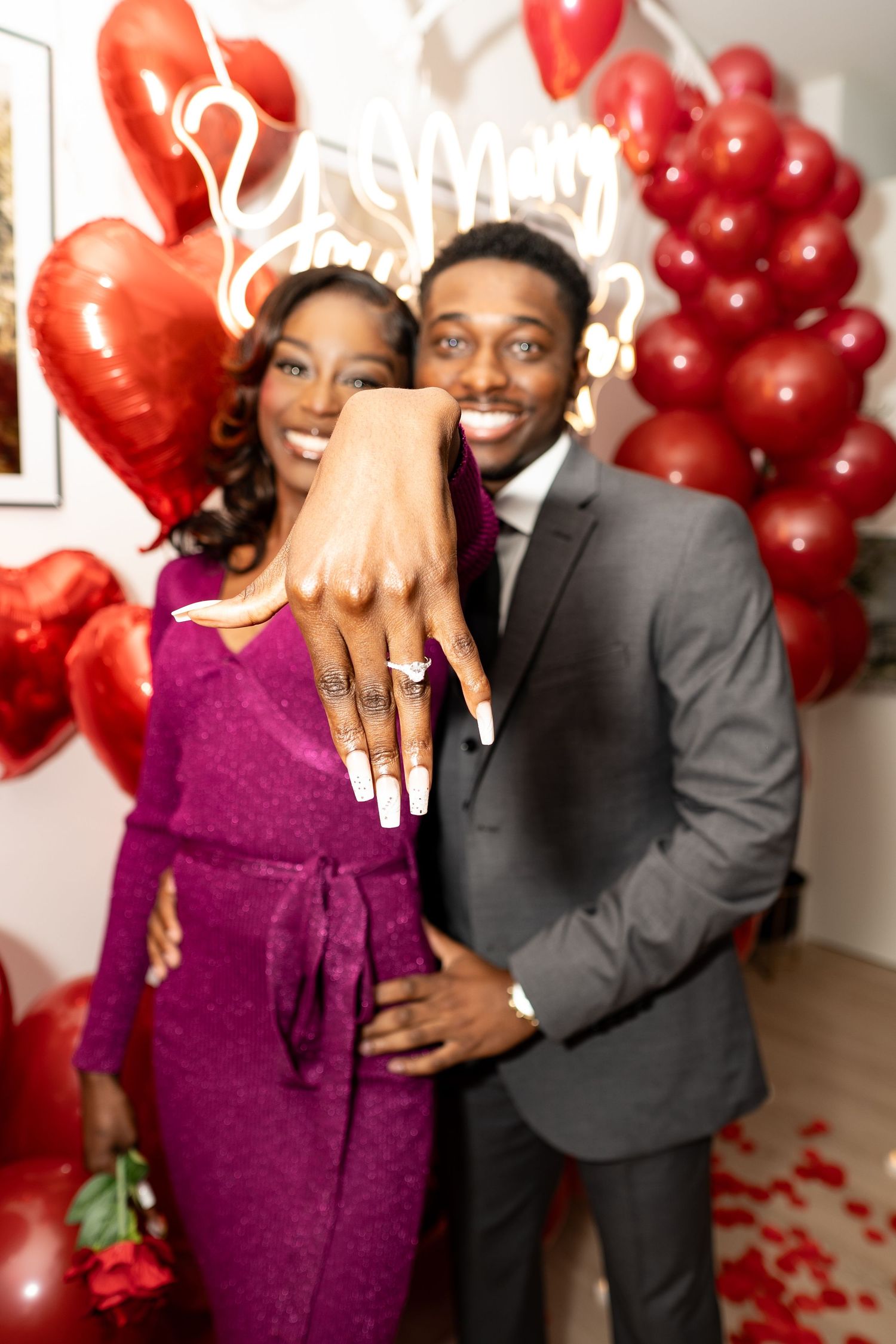 A couple poses together in front of red heart balloons and rose petals in a romantic Valentine's Day setting.