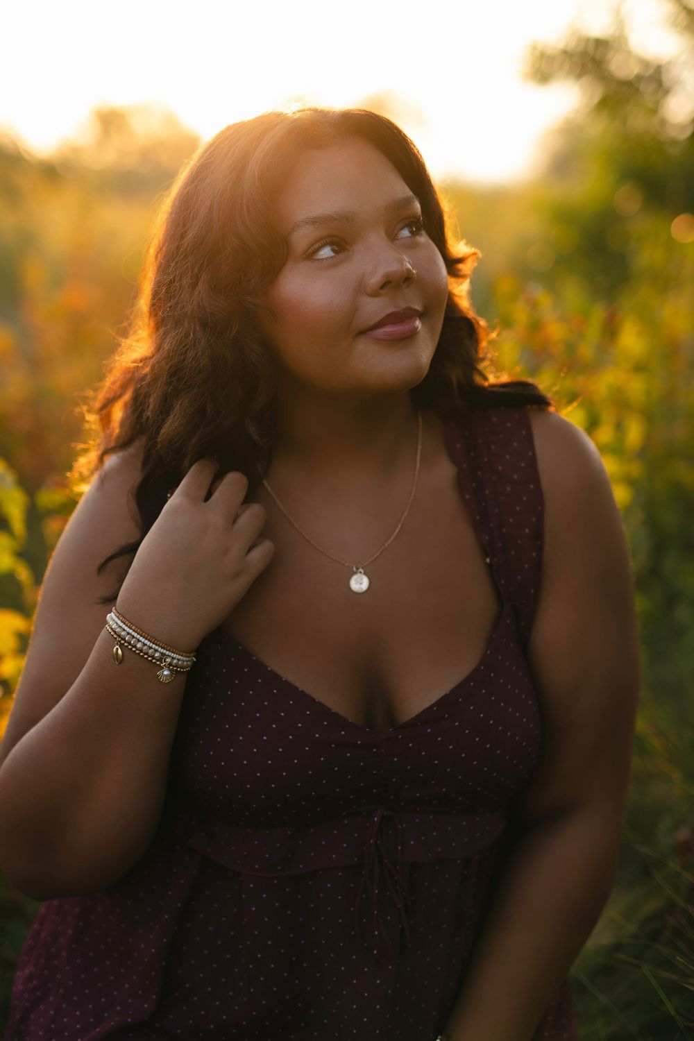 Person in a dark dress poses outdoors during golden hour with sunlit foliage in background.