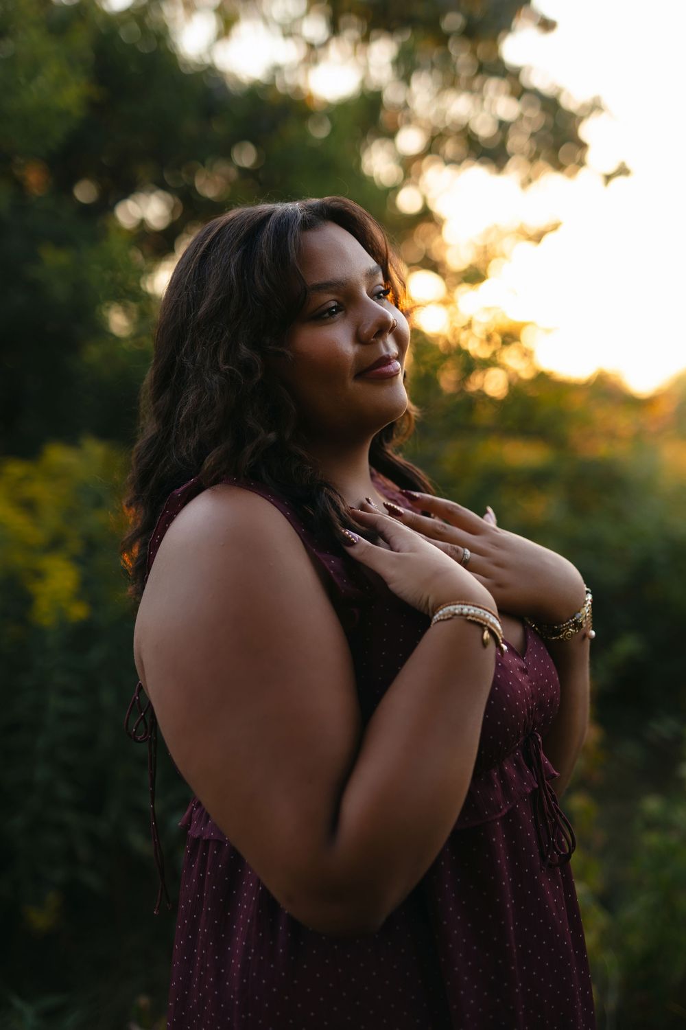 A figure in a burgundy lace dress poses gracefully during golden hour in a natural outdoor setting.