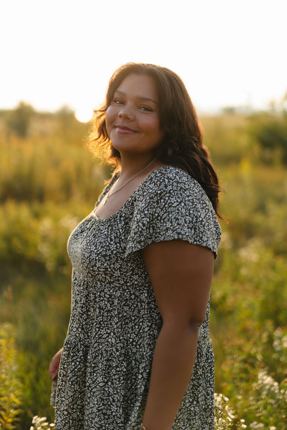 A person in a floral dress poses in a golden-lit meadow during sunset.