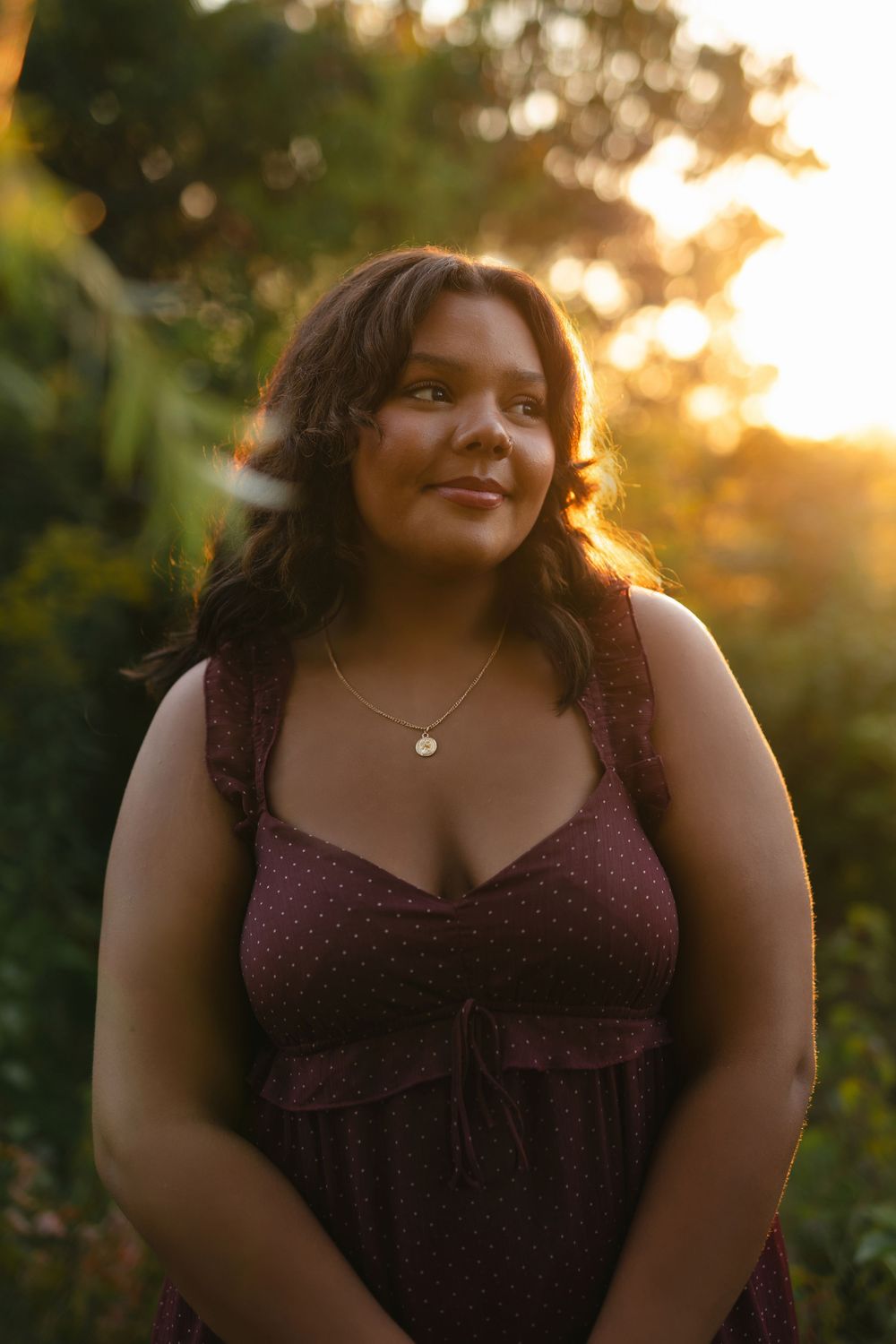A figure wearing a burgundy dress poses in a natural outdoor setting at golden hour.