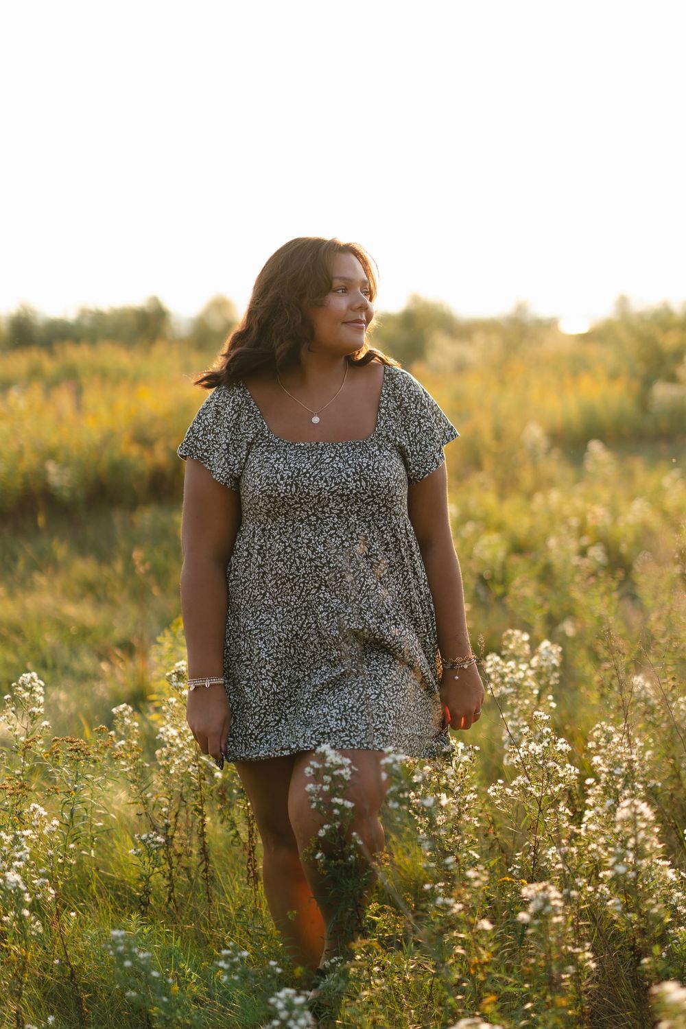 A person in a floral print dress stands in a golden field of wildflowers during sunset.