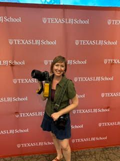 Woman with camera poses in front of Texas LBJ School branded red backdrop at an event.