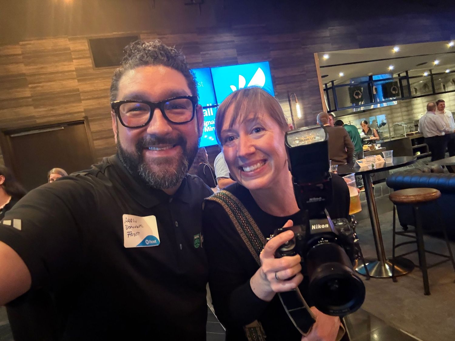 Two people smiling for a selfie at a bar event, woman holding a camera, man wearing a name tag.