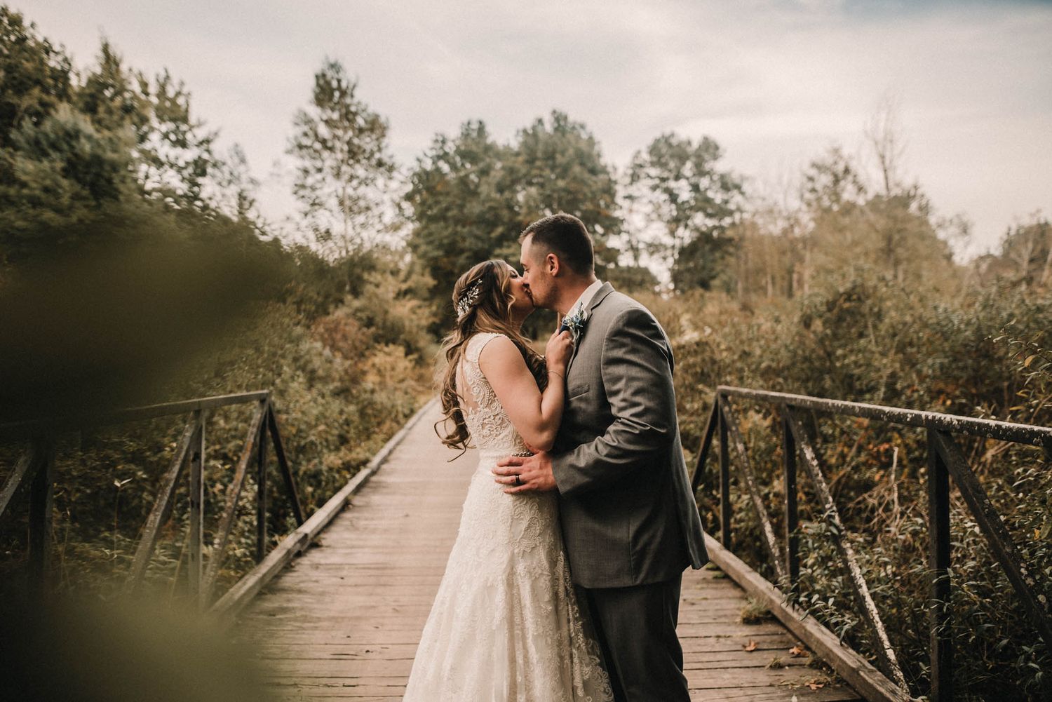 Romantic couple in wedding attire share tender moments on a rustic wooden bridge surrounded by autumn foliage.