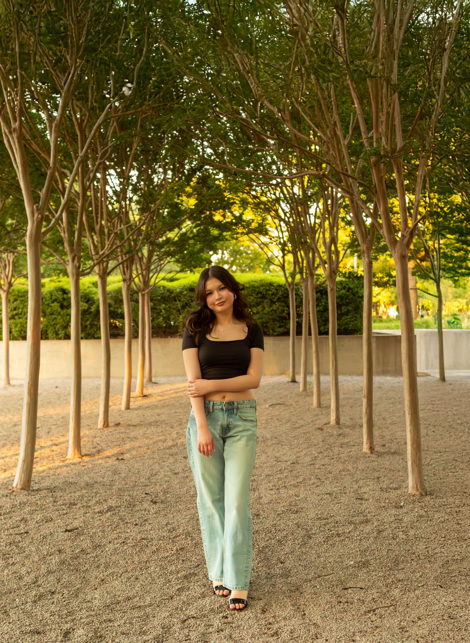 A person in black top and mint pants poses among a grove of young trees in a serene outdoor setting.