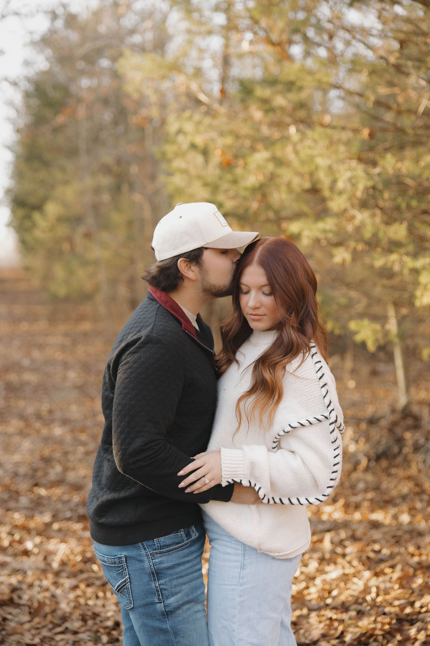 A romantic couple embraces in an autumn forest setting with fallen leaves and golden foliage.