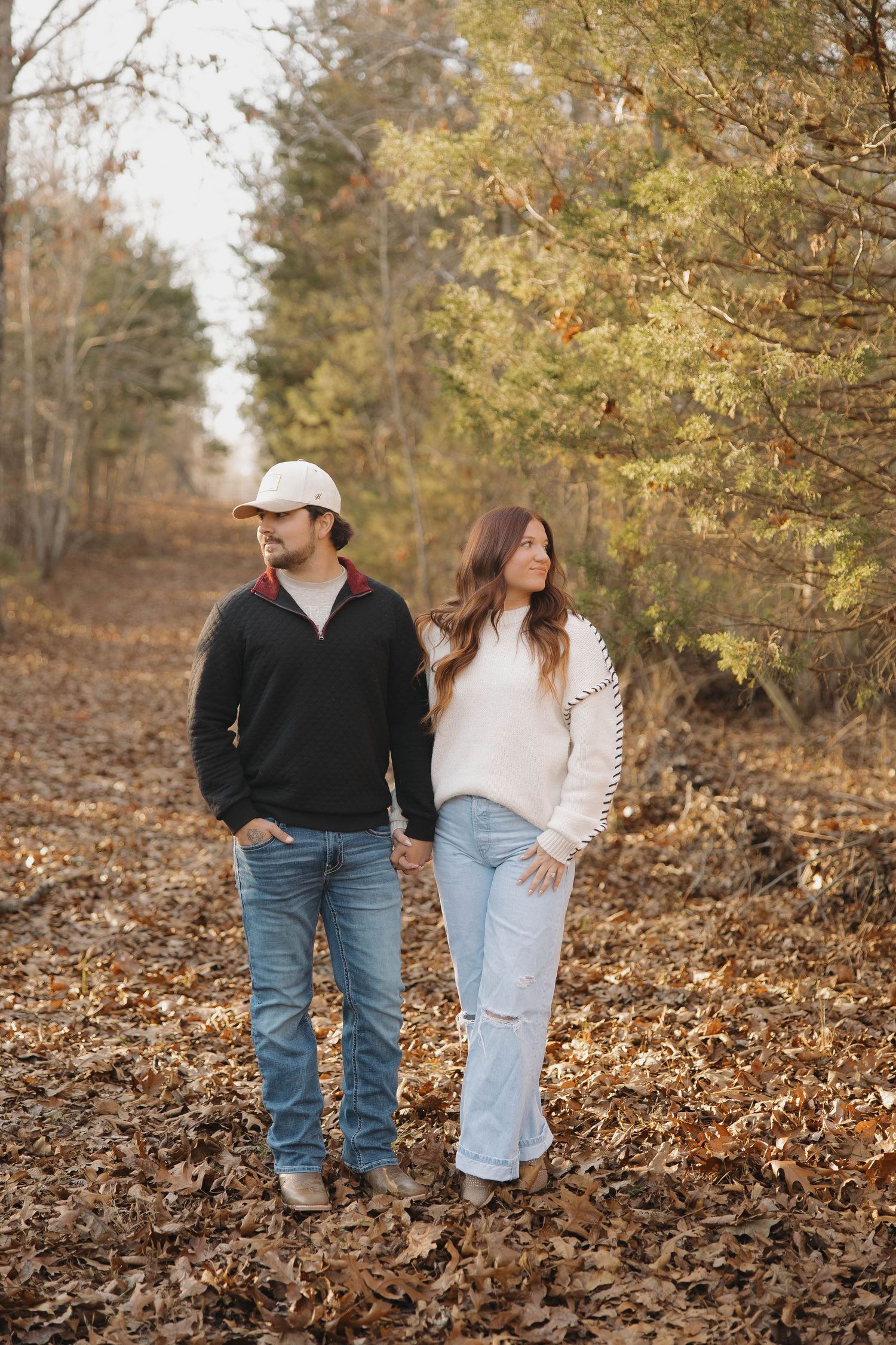 A couple in casual attire walking through autumn leaves in a wooded area on a fall day.