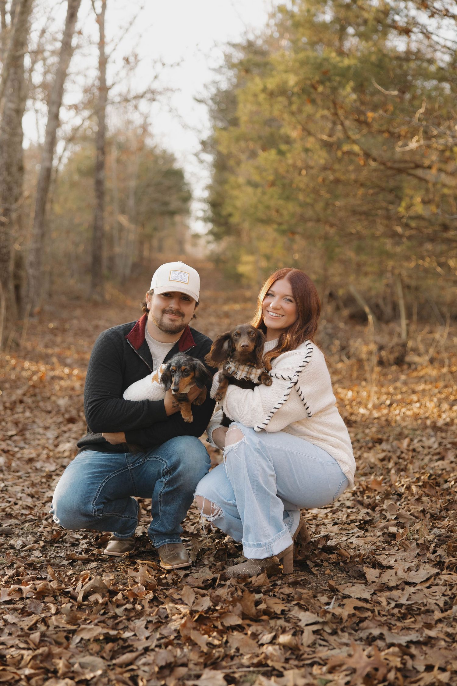 A couple sits together on fallen autumn leaves in a wooded area during a casual outdoor photoshoot.