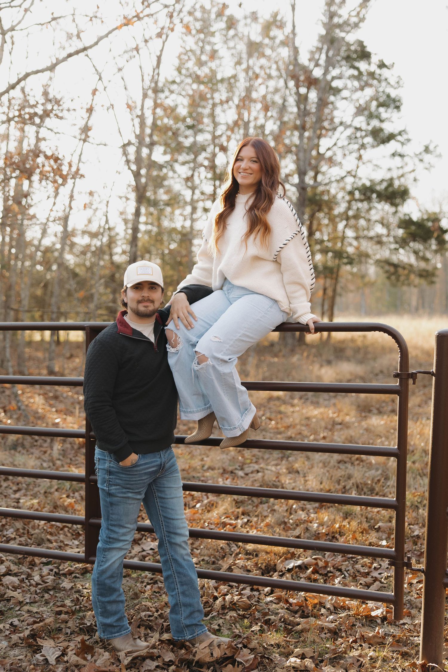 A couple playfully interacts while sitting on a rustic wooden fence during an autumn photoshoot in the woods.