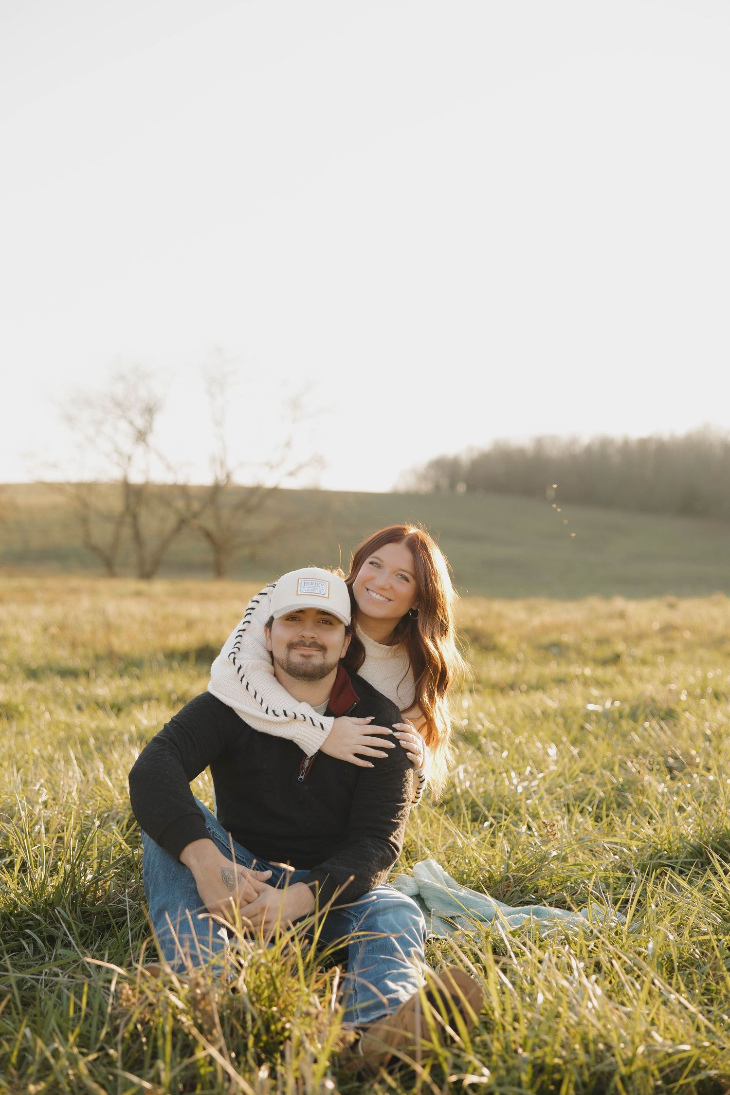A couple sits together in a sunlit meadow of tall grass during golden hour.