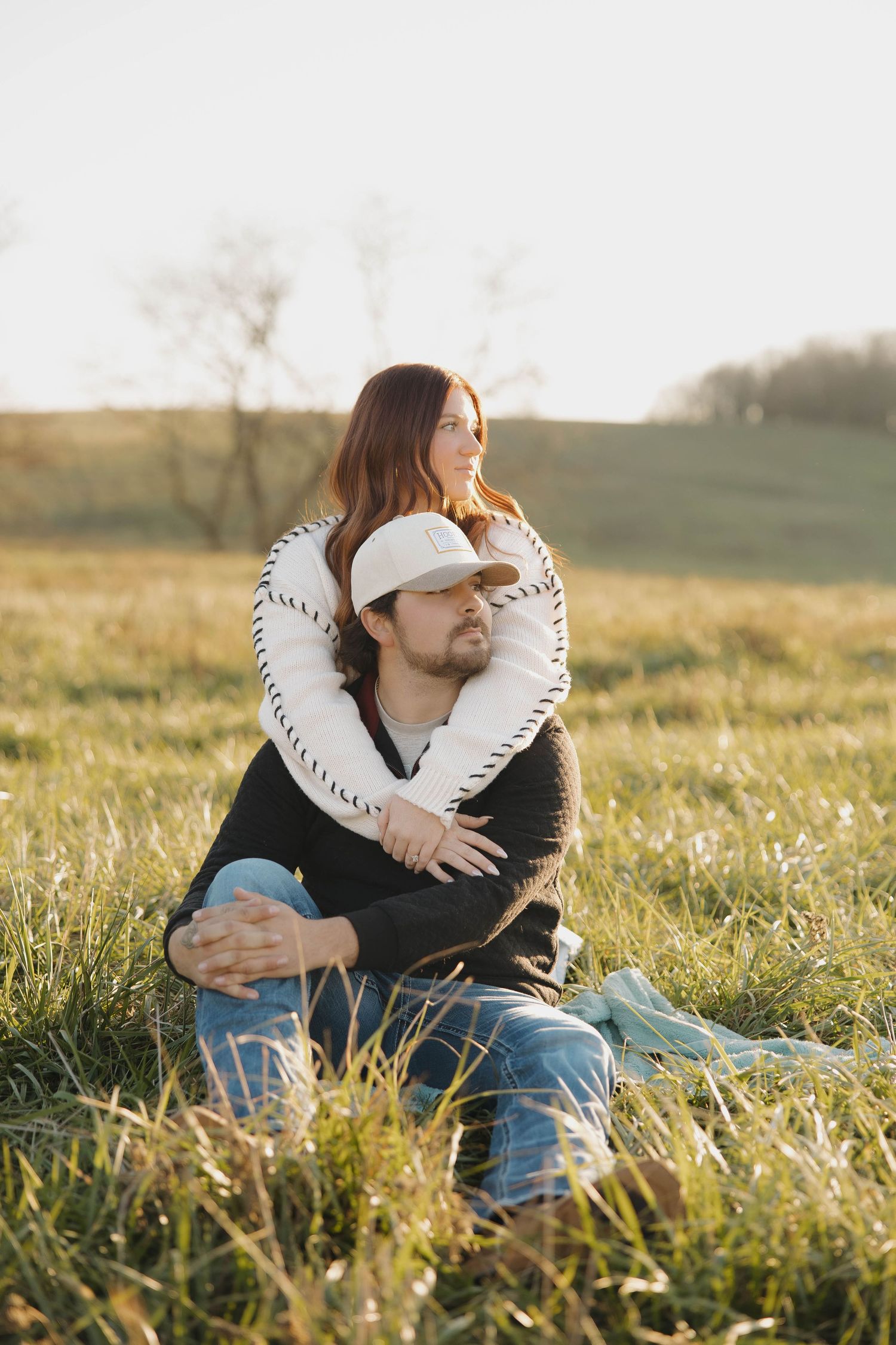 A couple sits together in a grassy meadow during golden hour, embracing while wearing casual clothing.