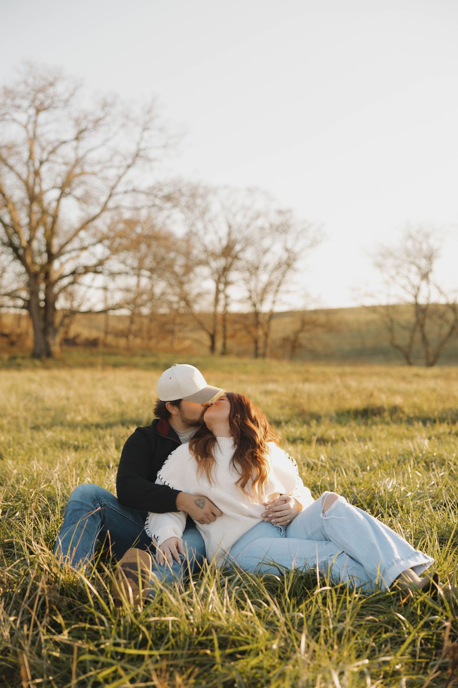 A couple sits together in a sunlit autumn field with tall grass and bare trees in the background.