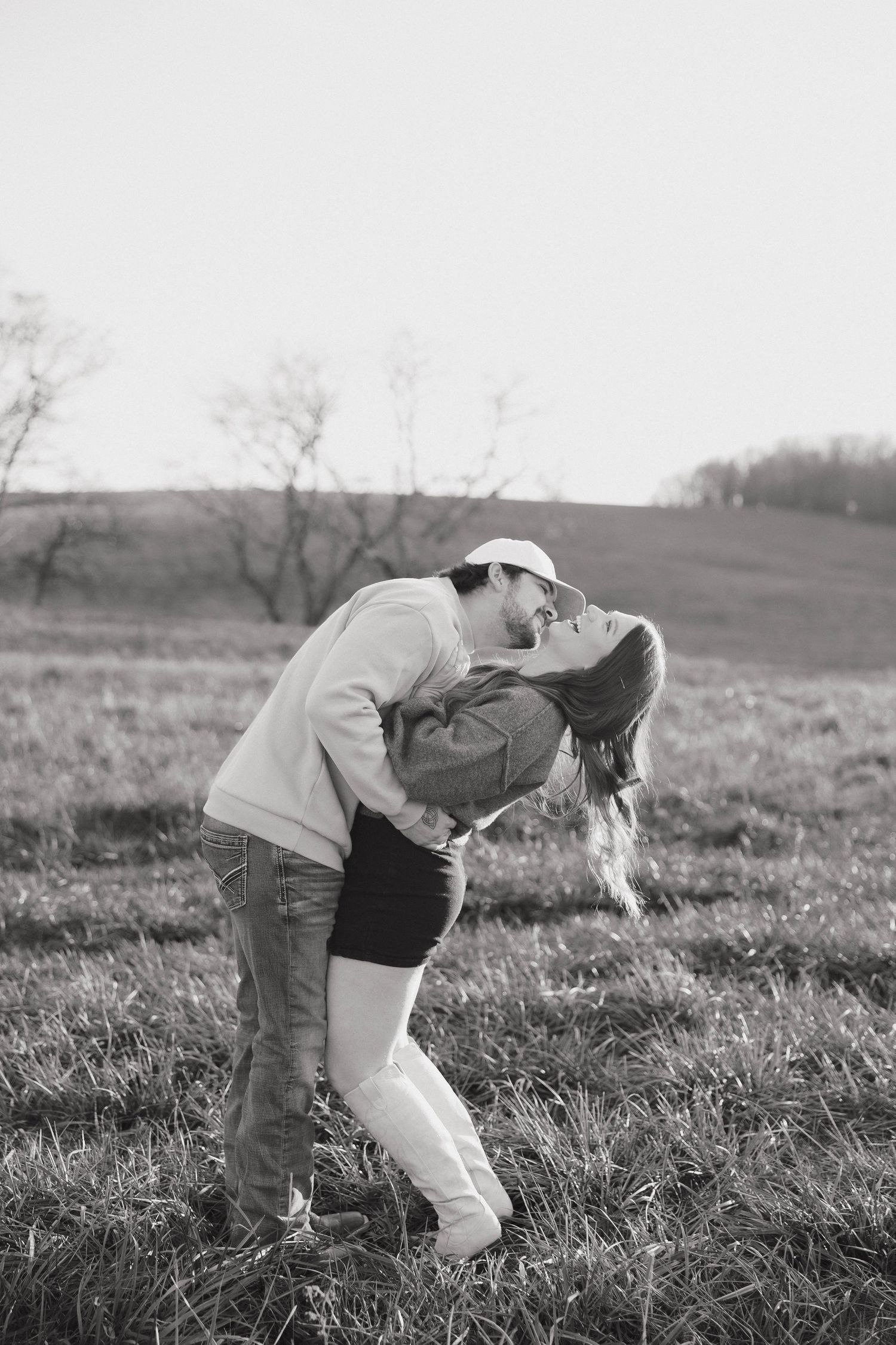 A romantic black and white photo of a couple embracing in a scenic field with bare trees in the background.