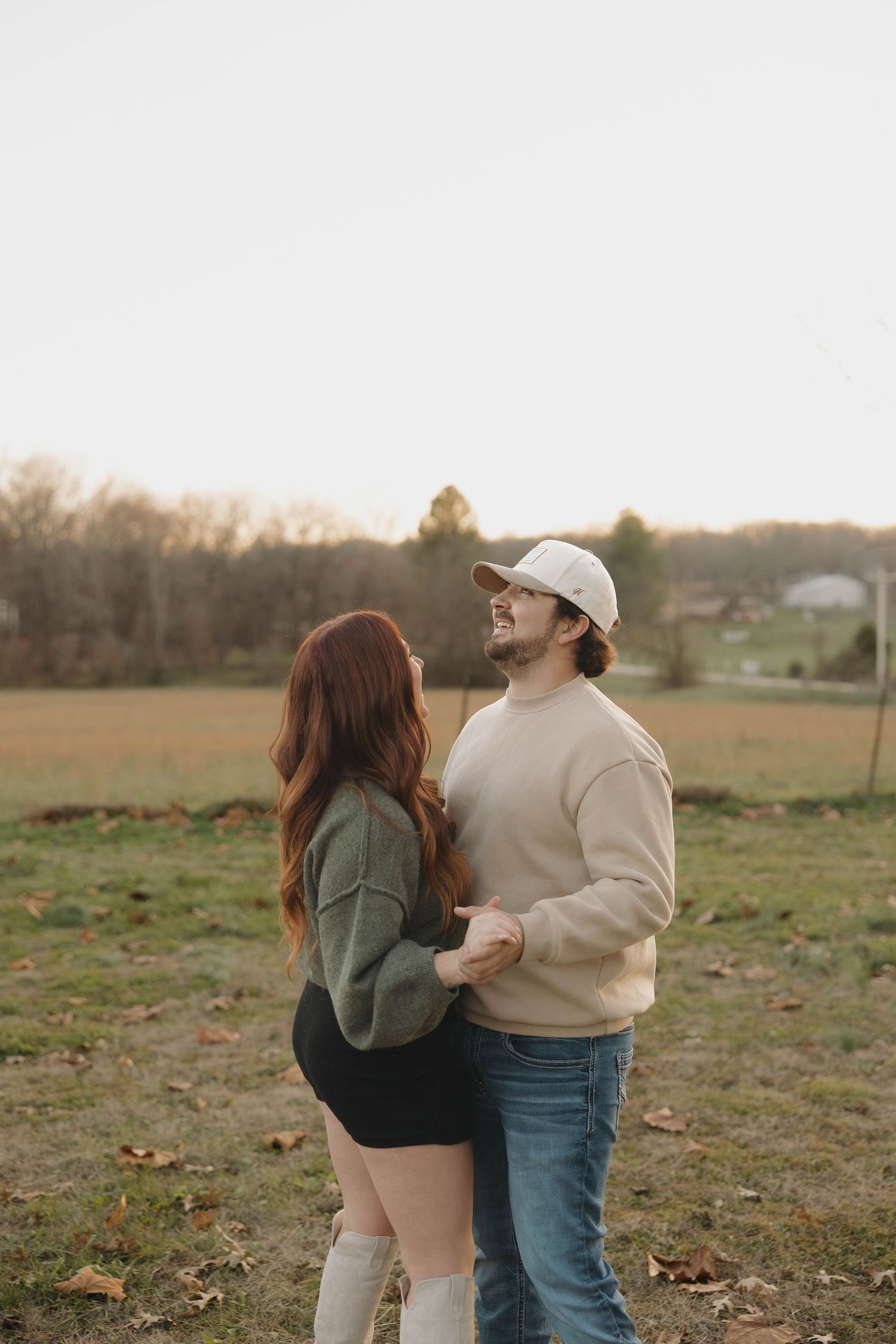 A couple in casual sweaters stands together in a rustic autumn field at sunset.