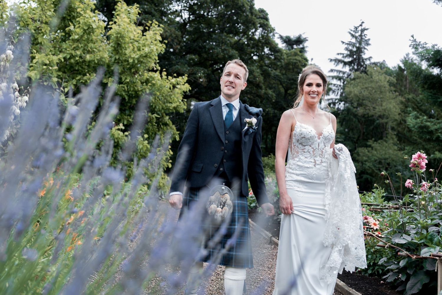 Newlyweds walk together along garden path with lavender flowers in foreground and trees in background.