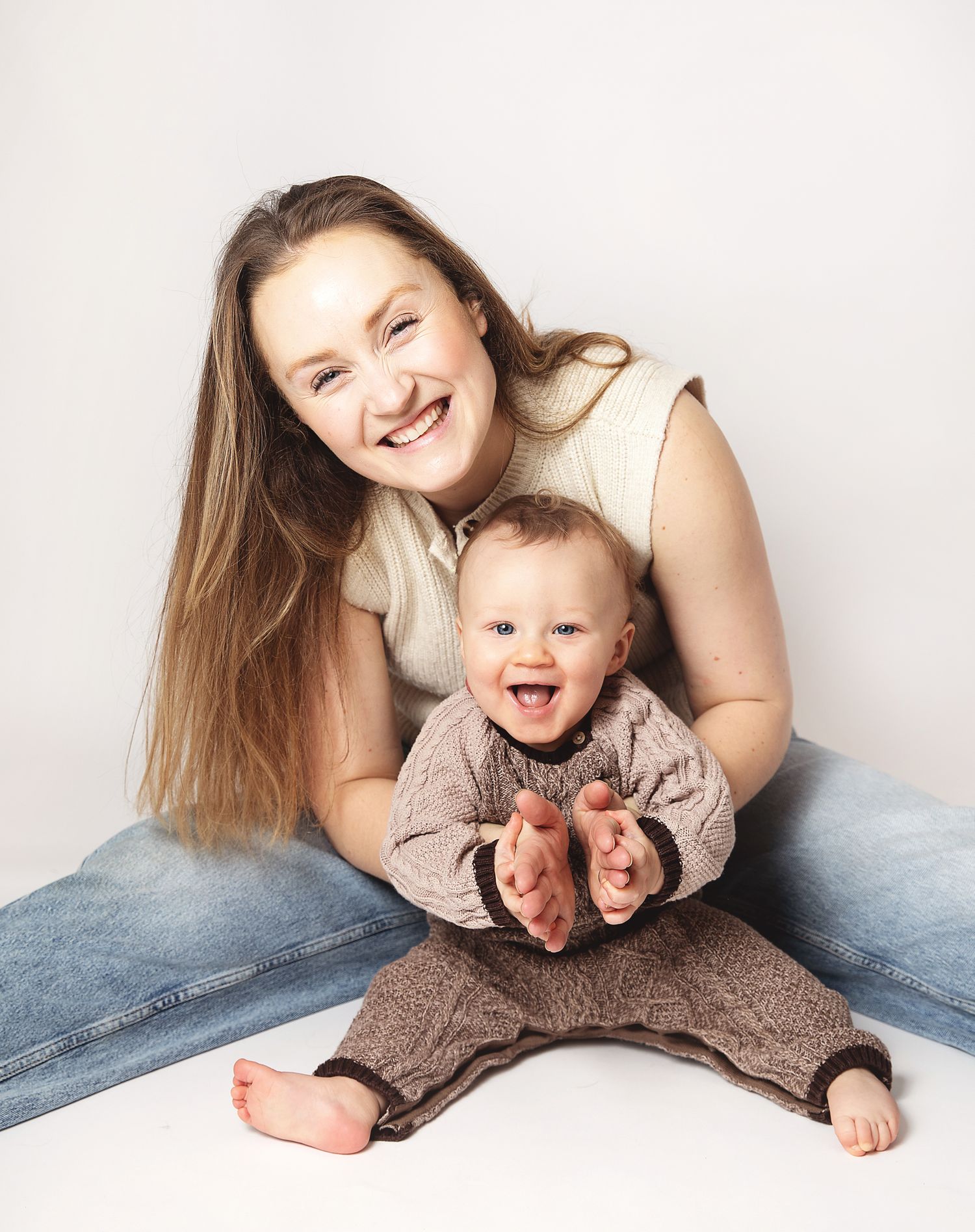 Mother and baby sitting together on white background wearing casual clothing while smiling joyfully. During a photoshoot in Sidcup