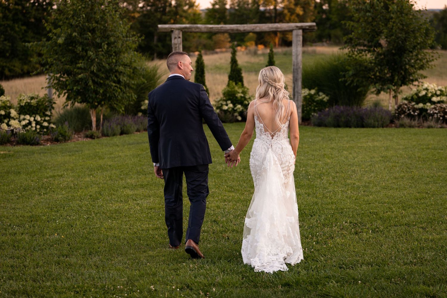 Couple in wedding attire walking together on green lawn at sunset.