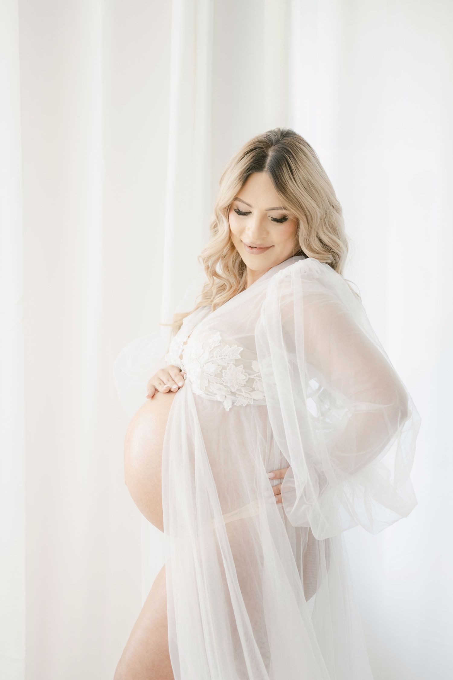 A glowing maternity shoot features a woman in sheer white lace robe sitting gracefully on a white bed with flowers nearby.