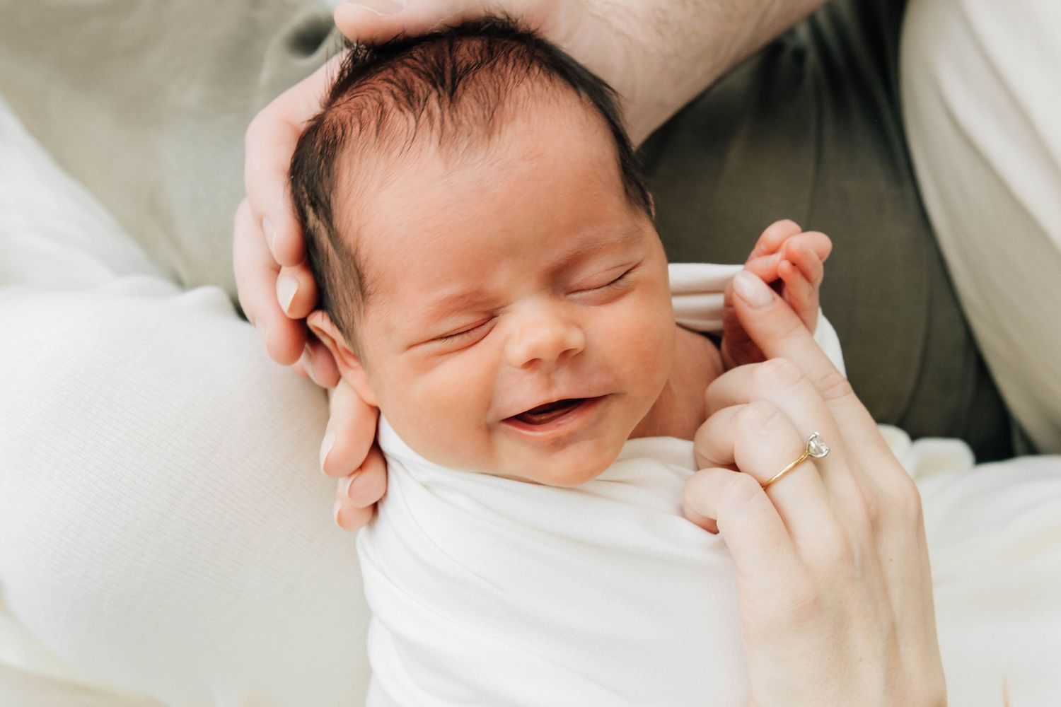 Smiling newborn baby swaddled in white with parents hands gently cradling their head.