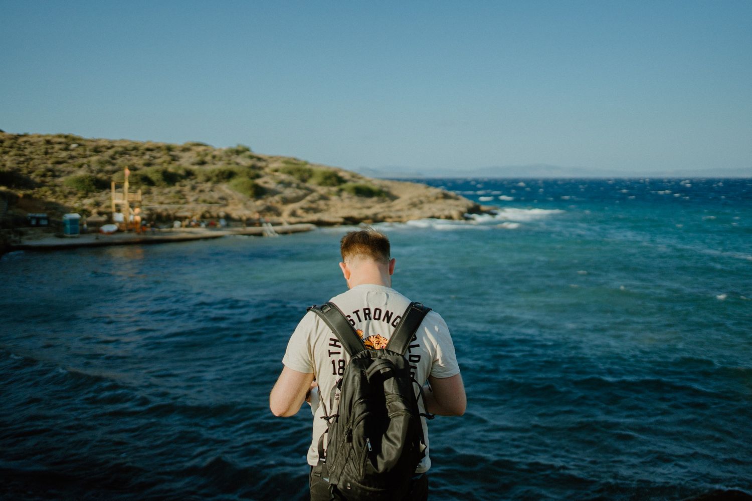 Person with backpack looking out at blue ocean waters and rocky coastline on sunny day.
