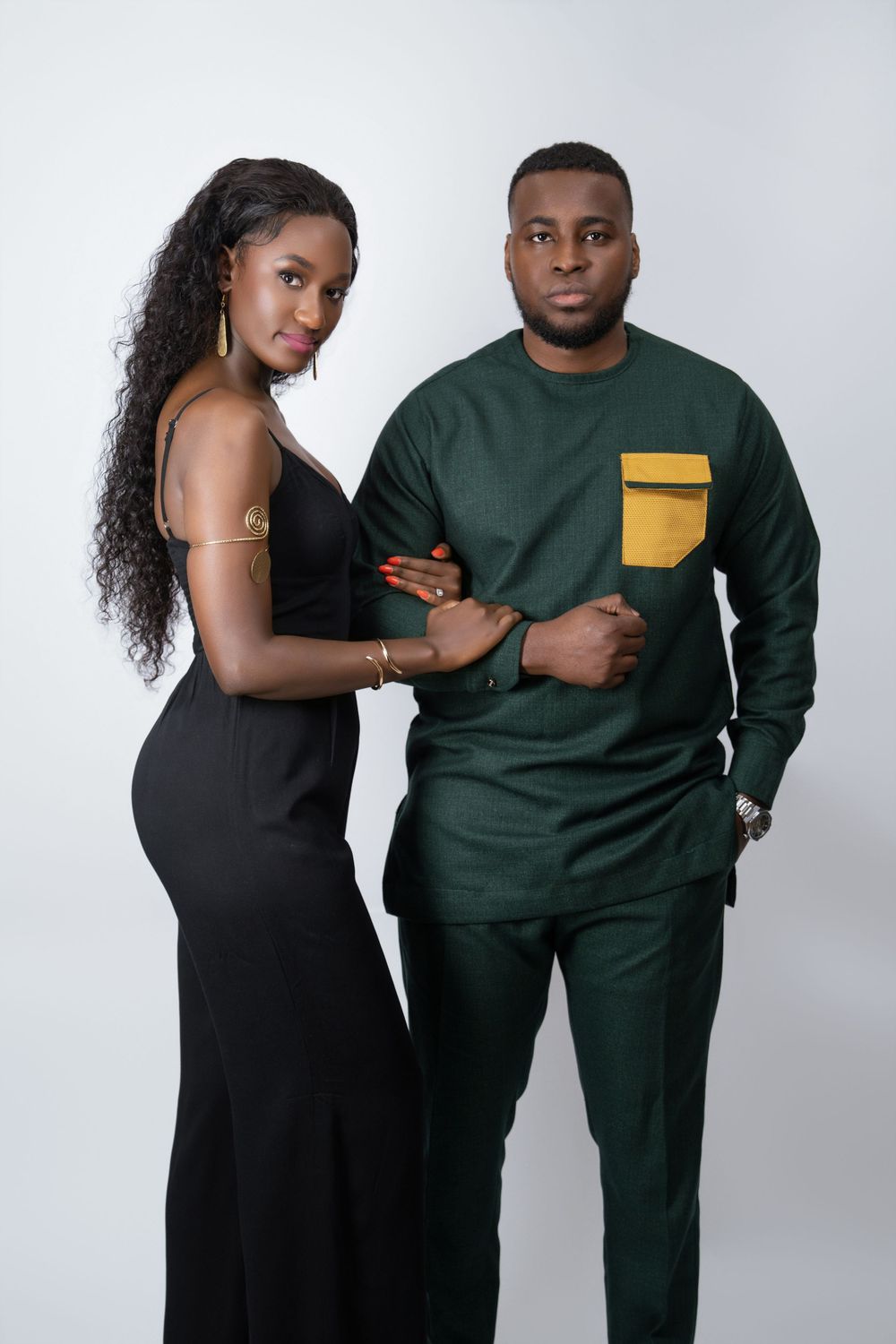 Couple in formal attire posing against white backdrop, wearing black dress and green traditional outfit.