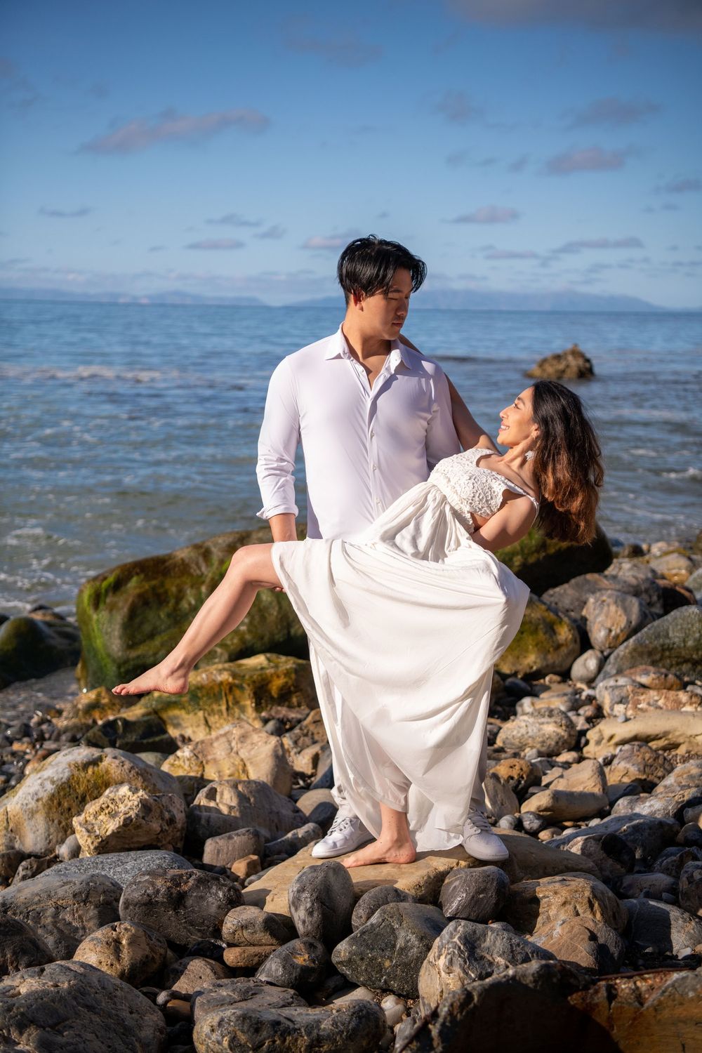 Romantic couple poses on rocky beach shore with ocean backdrop.