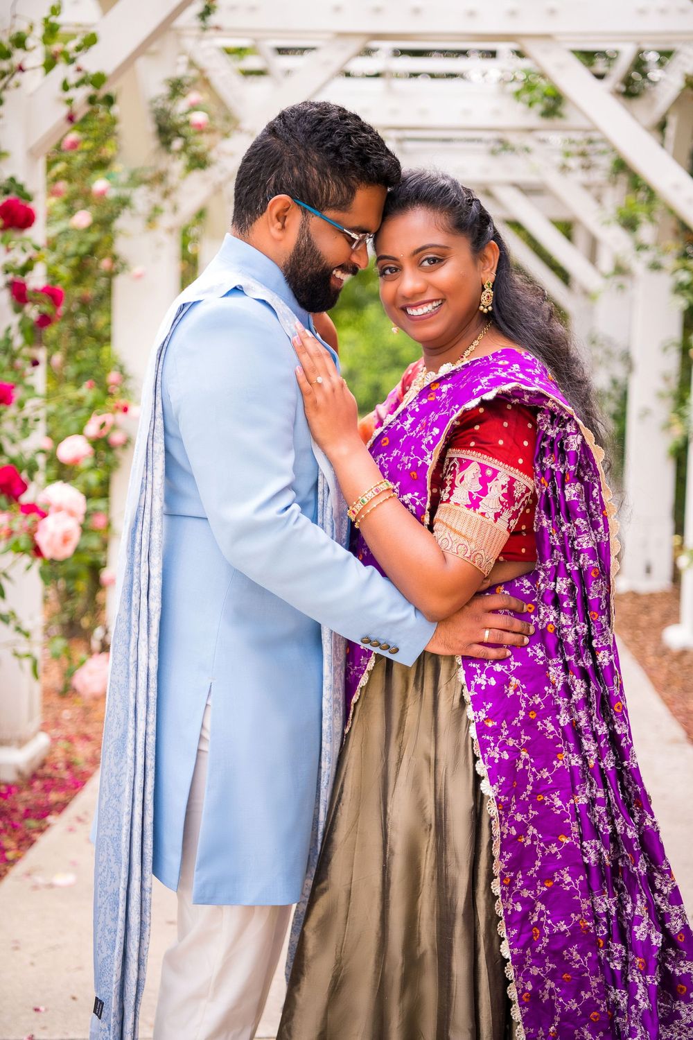 Couple in traditional Indian attire embrace in front of rose-covered garden trellis.