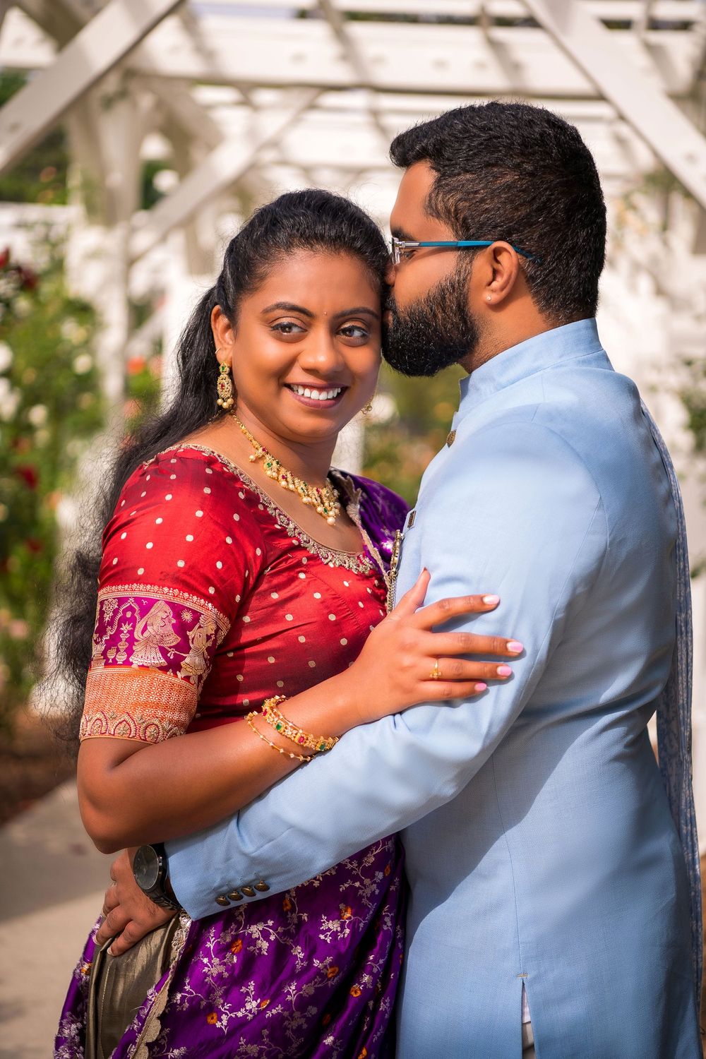 A romantic couple in traditional Indian attire embrace under a pergola in a garden setting.
