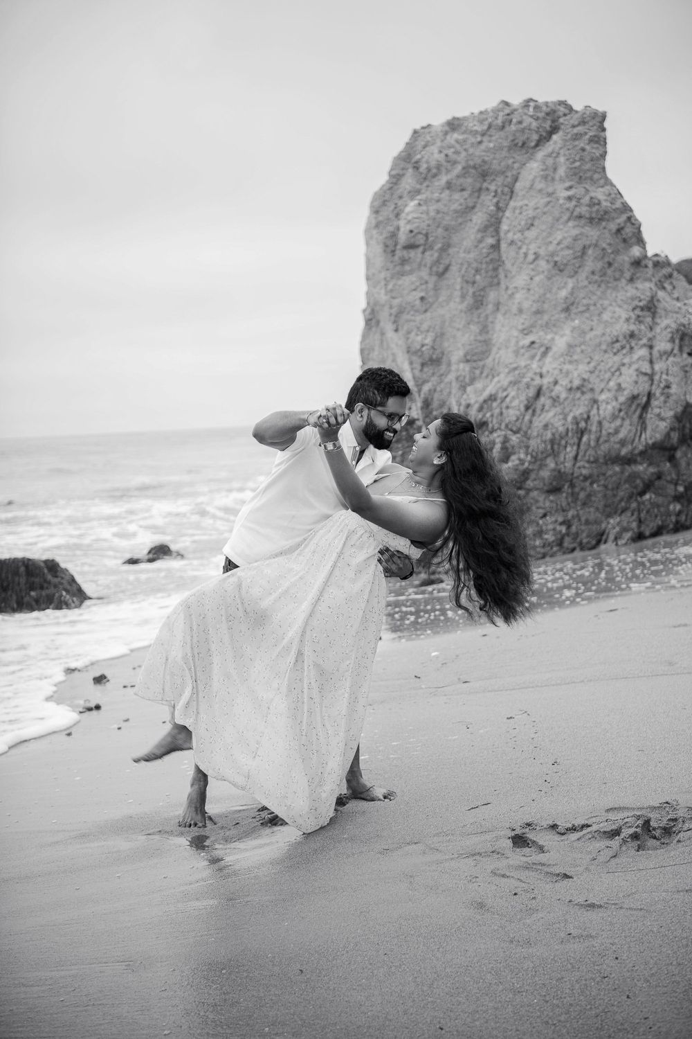 A romantic black and white beach photo with dramatic coastal rocks in the background.