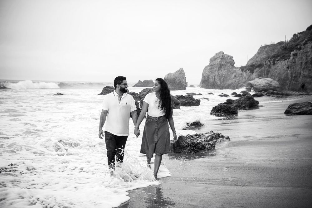 A couple walking together on a beach with crashing waves and rocky coastline.