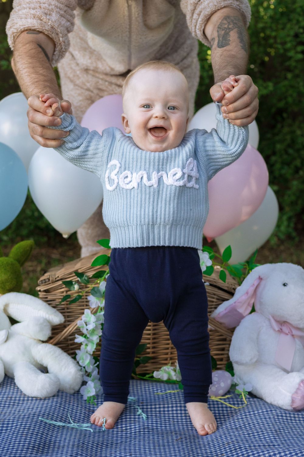 Baby boy standing up holding onto someone's hands standing behind them, wearing a blue jumper with 'BENNETT' spelled out on it