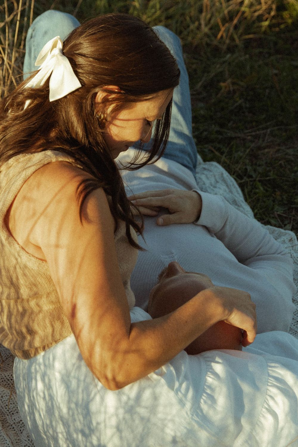 Pregnant woman in a white dress sitting in sunlit grass cradling her baby bump.