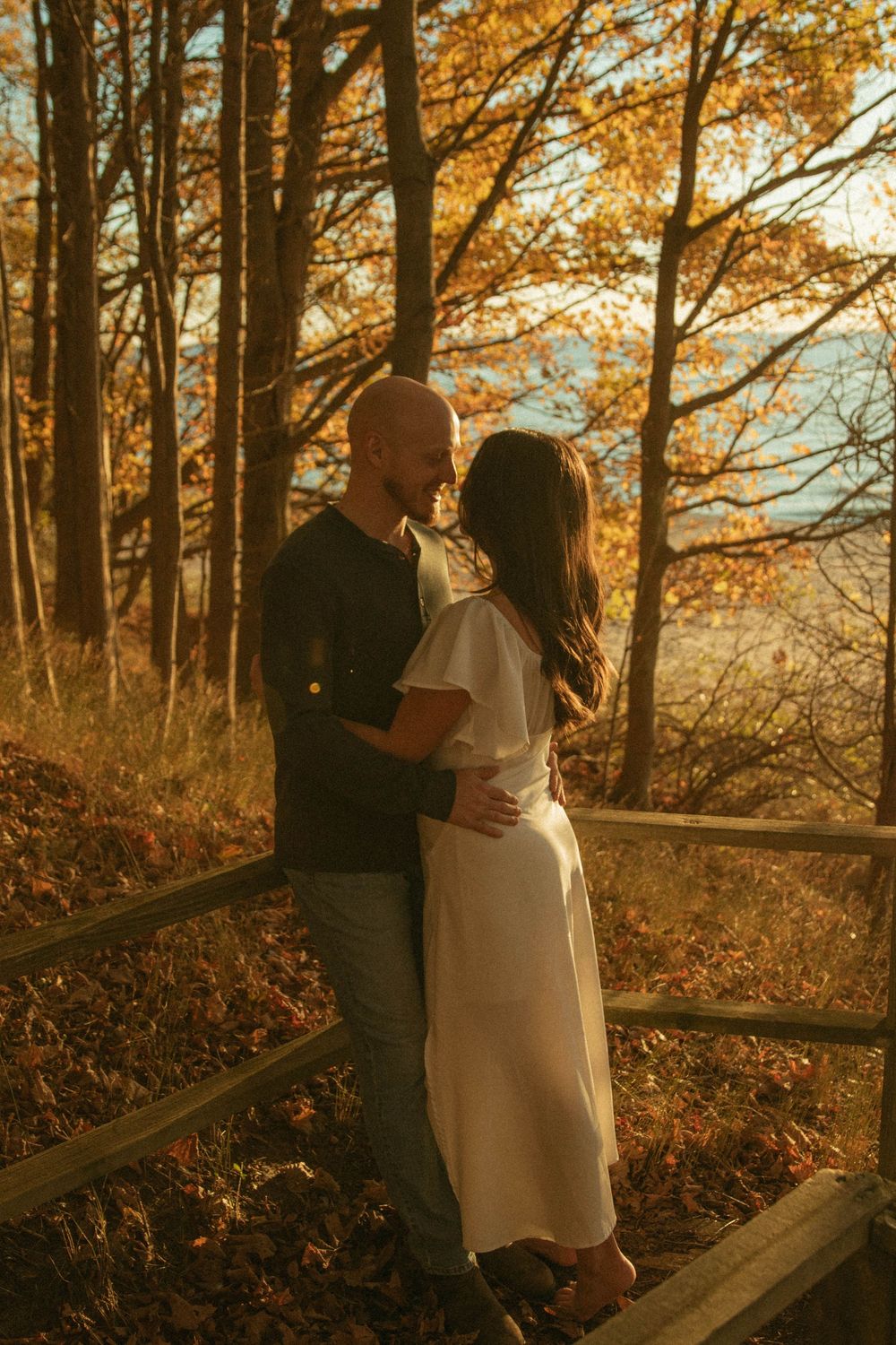 Couple embracing near a wooden fence surrounded by autumn foliage in warm sunlight.